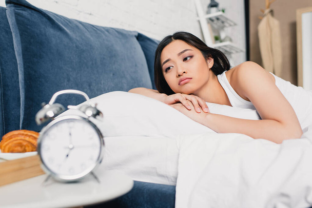 Szelektív fókusz a szomorú ázsiai lány feküdt az ágyon közelében ébresztőóra és reggeli az éjjeliszekrényen  - Fotó, kép