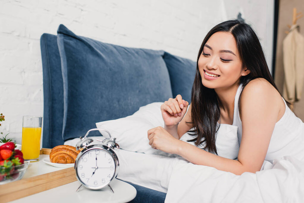 Selective focus of smiling asian girl lying on bed near breakfast and alarm clock on bedside table  - Valokuva, kuva