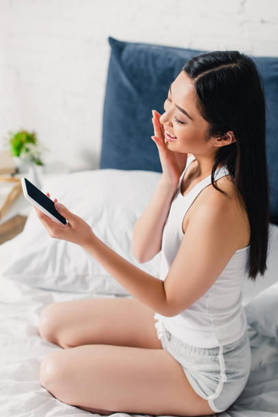 Sorrindo asiático mulher segurando smartphone com tela em branco na cama de manhã
  - Foto, Imagem