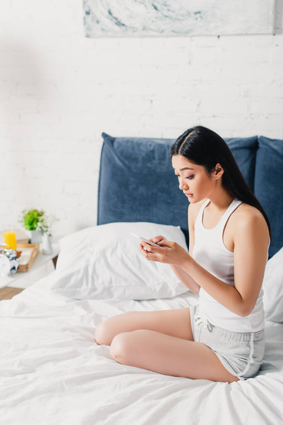 Surprised asian girl using smartphone while sitting on bed at home  - Fotoğraf, Görsel