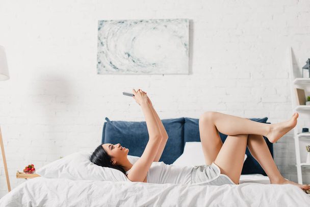 Oldalnézet pozitív ázsiai lány használ okostelefon, miközben fekszik az ágyon otthon  - Fotó, kép