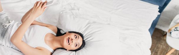 Panoramic shot of positive asian girl holding smartphone and looking away on bed  - Photo, Image