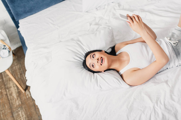 Top view of smiling asian woman holding smartphone and looking away on bed at morning  - Foto, Imagen