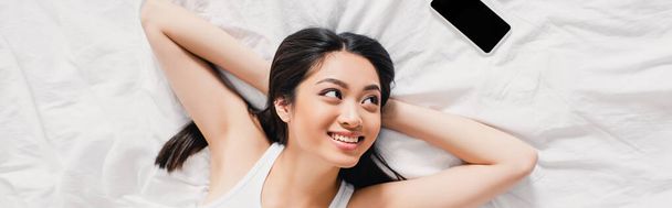 Horizontal image of smiling asian woman looking at smartphone with blank screen on bed  - 写真・画像
