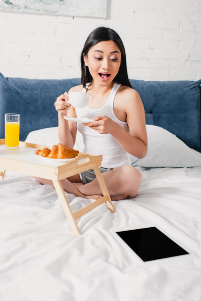 Excited asian woman looking at digital tablet while drinking coffee on bed  - Foto, Imagen