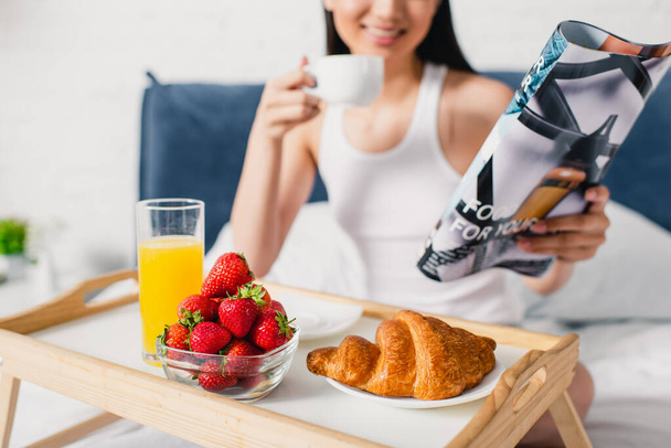 A croissant, az eper és a narancslé szelektív fókusza a reggelizőtálcára és a mosolygó lányra, aki kávét és magazint tart az ágyon  - Fotó, kép