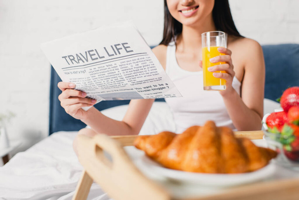 Ausgeschnittene Ansicht einer lächelnden Frau, die morgens eine Zeitung mit Reiseaufdruck und einem Glas Orangensaft auf dem Bett hält  - Foto, Bild