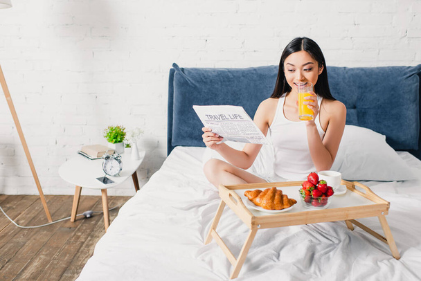 enfoque selectivo de sonriente chica asiática leyendo periódico durante el desayuno con jugo de naranja y croissant en el dormitorio - Foto, imagen