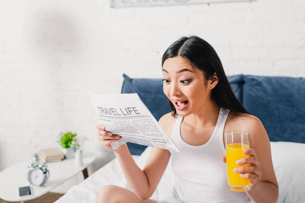 Opgewonden aziatische vrouw leest krant met reisleven belettering en houden glas sinaasappelsap op bed  - Foto, afbeelding