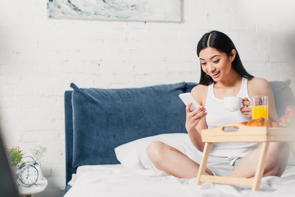 Szelektív fókusz mosolygós ázsiai lány okostelefon és gazdaság kávéscsésze közelében reggeli tálcán az ágyon  - Fotó, kép