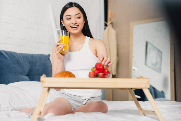 Selective focus of cheerful asian girl holding glass of orange juice near breakfast with strawberries on bed  - 写真・画像