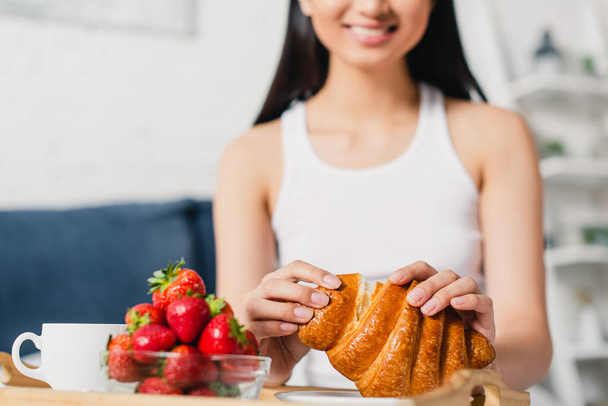 Selective focus of smiling woman holding croissant near strawberries and coffee in bedroom - Foto, Imagem