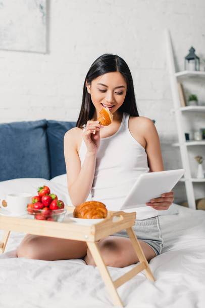 Selective focus of asian girl eating croissant and using digital tablet during breakfast on bed  - Photo, Image