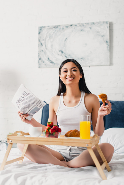 Selective focus of asian girl smiling at camera while holding newspaper and croissant on bed  - Photo, Image