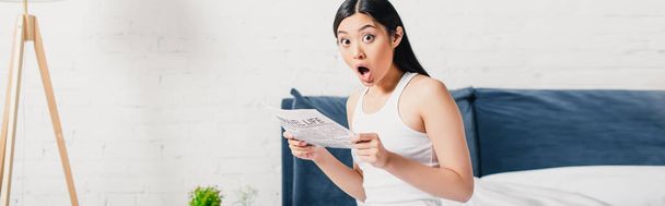 Horizontal crop of shocked asian woman holding newspaper on bed  - Photo, Image