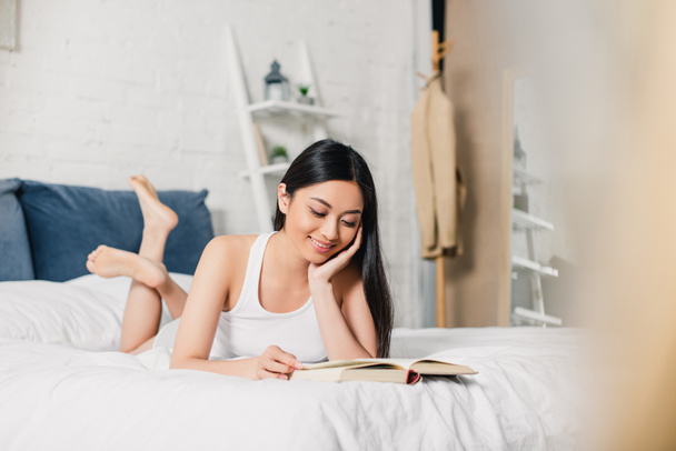 Выборочный фокус улыбающейся азиатской девушки, читающей книгу, лежащей дома на кровати  - Фото, изображение