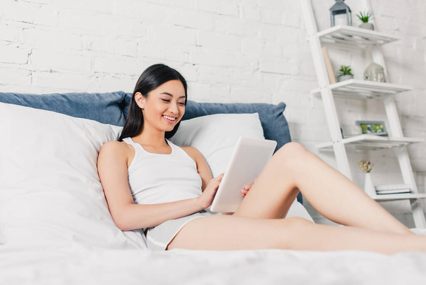 Selective focus of beautiful asian woman smiling and using digital tablet on bed  - Фото, зображення