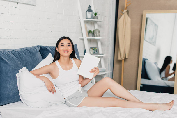 Bella donna asiatica che tiene tablet digitale e sorride alla fotocamera sul letto a casa  - Foto, immagini