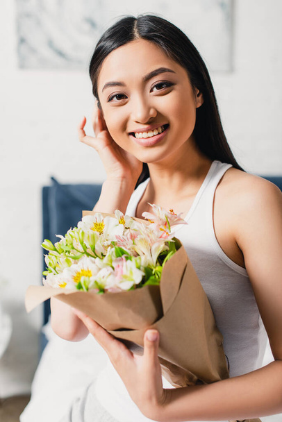 Valikoiva painopiste aasialainen nainen hymyilee kameralle ja tilalla kukkakimppu makuuhuoneessa - Valokuva, kuva