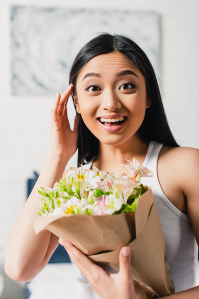Selektiver Fokus fröhlicher asiatischer Mädchen mit Blumenstrauß und Blick in die Kamera zu Hause  - Foto, Bild
