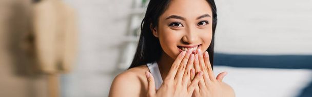 Panoramic shot of positive asian woman with hands near mouth looking at camera at home  - Foto, Imagem