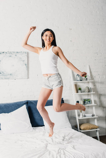 Cheerful asian woman in pajamas jumping on bed  - 写真・画像