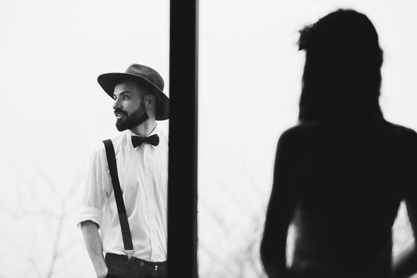 portrait of a stylish wedding couple: Young bearded man in hat and brunette bride in elegant gown wearing a wreath braided in hairstyle - Foto, Imagen