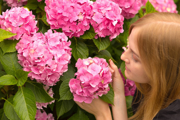 Gyönyörű, fiatal szőke nő, virágokkal az arca mellett. Rózsaszín Hydrangea makrophylla virágzás nyáron a kertben - Fotó, kép