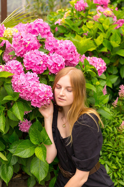 Beautiful young blonde woman with flowers near face. Pink Hydrangea macrophylla blooming in summer in garden - Photo, Image