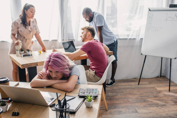 Foco seletivo de empresária chateada olhando para laptop enquanto colegas multiétnicos trabalhando em segundo plano
  - Foto, Imagem
