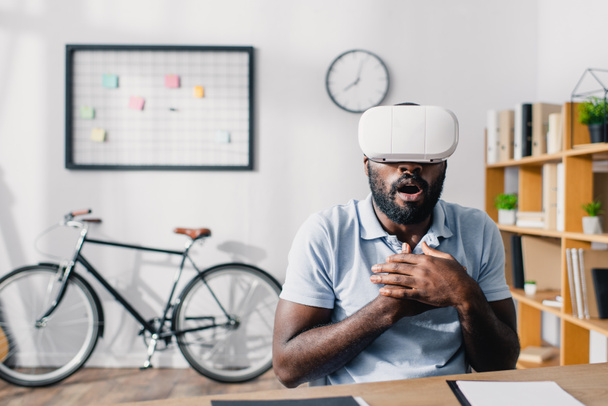 Selektiver Fokus aufgeregter afrikanisch-amerikanischer Geschäftsmann mit Virtual-Reality-Headset am Tisch im Büro  - Foto, Bild