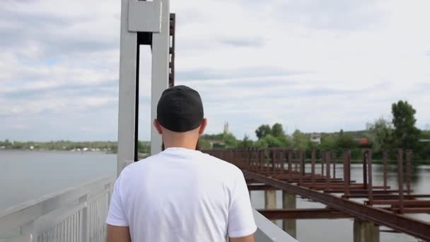 A young guy walks on a narrow bridge and looks back towards the camera - Materiał filmowy, wideo