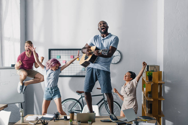 Enfoque selectivo del empresario afroamericano tocando la guitarra acústica mientras colegas multiétnicos bailan en la oficina  - Foto, Imagen