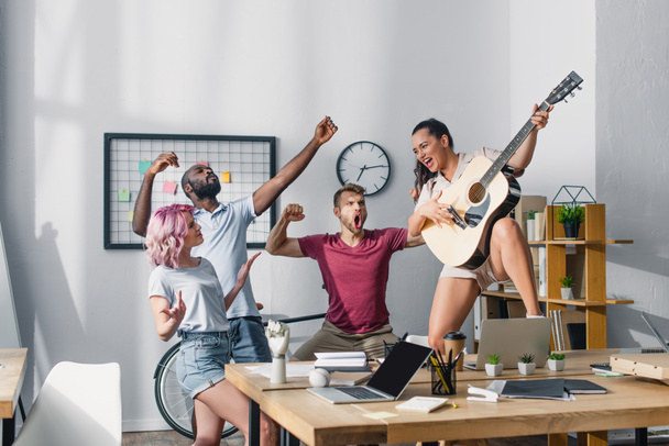 Επιλεκτική εστίαση των επιχειρηματιών που παίζουν ακουστική κιθάρα κοντά σε πολυπολιτισμικούς συναδέλφους τραγουδώντας και χορεύοντας στο γραφείο  - Φωτογραφία, εικόνα