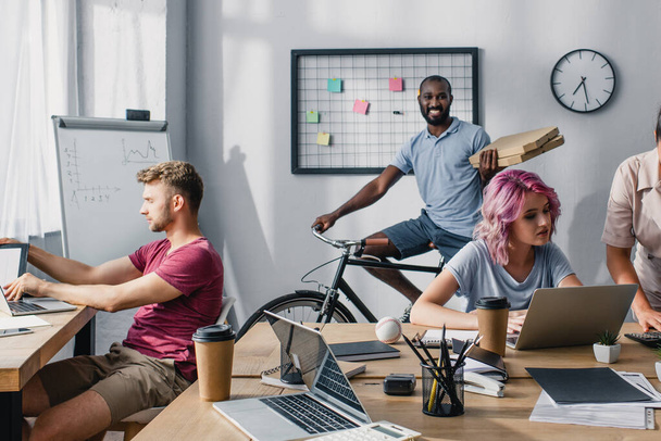 Concentration sélective de l'homme d'affaires afro-américain sur le vélo tenant des boîtes à pizza près de collègues multiculturels travaillant dans le bureau  - Photo, image
