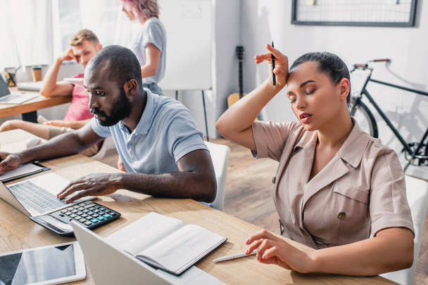 Selectieve focus van vermoeide zakenvrouw houden pen in de buurt van Afrikaanse Amerikaanse collega met behulp van laptop in het kantoor  - Foto, afbeelding