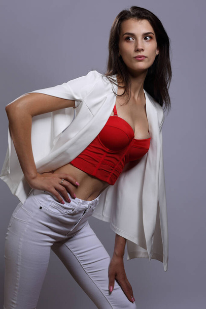 Portrait of a beautiful girl in a red bustier and white jeans on a gray background - Фото, зображення