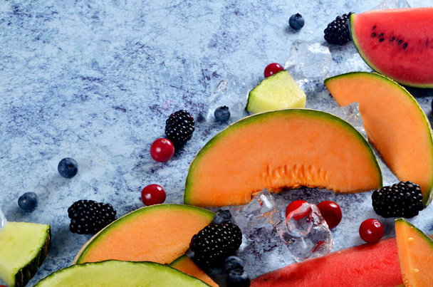 Verse gesneden meloen, watermeloen, bessen en ijsblokjes op een blauwe stenen ondergrond. Kopieer ruimte en tekstgebied - Foto, afbeelding