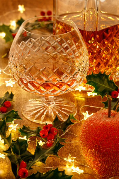 Kristallschnaps-Dekanter und ein Glas Brandy mit Weihnachtsbeleuchtung und Stechpalmen, auf einem Holztisch - Foto, Bild