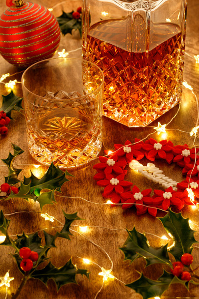 Κρυστάλλινο ποτήρι ουίσκι και κρυστάλλινη καράφα ουίσκι με χριστουγεννιάτικα φώτα και κόκκινο χριστουγεννιάτικο στεφάνι σε ξύλινο τραπέζι - Φωτογραφία, εικόνα