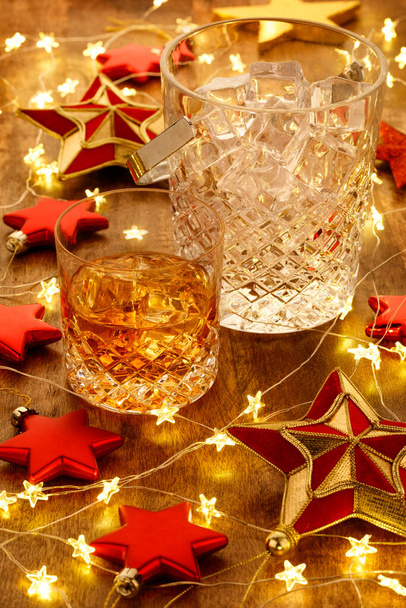 Crystal whisky ijsemmer en een glas whisky met kerstverlichting en rode sterren decoratie, op een houten tafel - Foto, afbeelding
