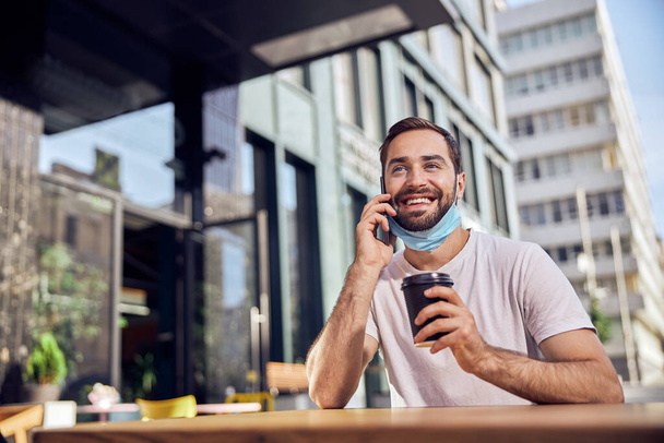 Man with a lowered mask is drinking coffee and talking on the smartphone  - Photo, Image