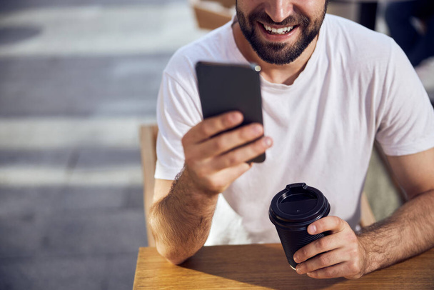 Parte de la cara de hombre sentado en la mesa en la cafetería con café y mirando al teléfono inteligente de cerca - Foto, Imagen