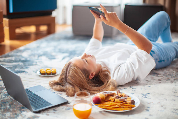 Teen girl lying on floor with laptop and phone scrolling the feed in social networks. Cozy home environment - Foto, afbeelding