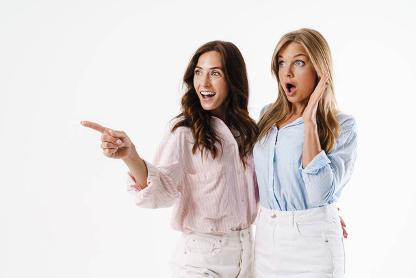 Image of excited beautiful two women pointing finger aside while hugging isolated over white background - Фото, изображение