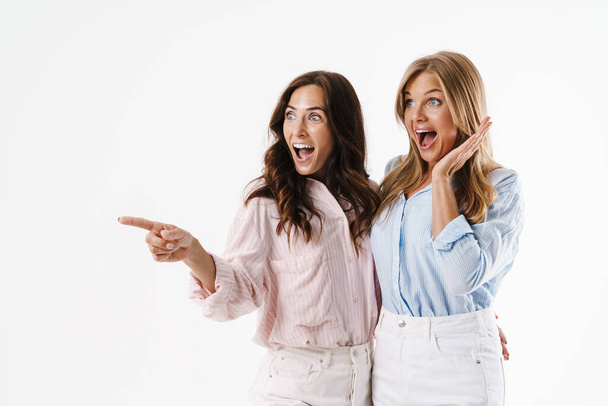 Image of surprised beautiful two women pointing finger aside while hugging isolated over white background - Foto, Imagem
