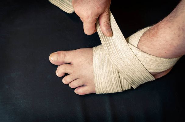 Middle-aged Caucasian man produces an independent bandage of his dislocated leg. He puts an elastic bandage over his swollen shin. First aid in sprains and dislocations. Bandage stabilizes ankle joint. - Photo, Image