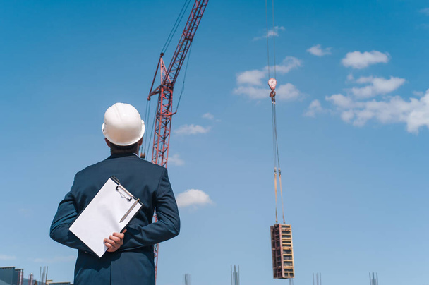 The chief in a jacket and a construction helmet with documents in his hands, against the background of a tower crane and the sky with clouds. House construction inspection - Fotó, kép