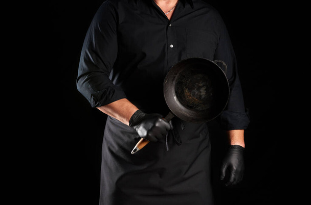 macho cozinheiro em uniforme preto e luvas de látex detém uma rodada vazia de ferro fundido preto vintage na frente dele, chave baixa
 - Foto, Imagem