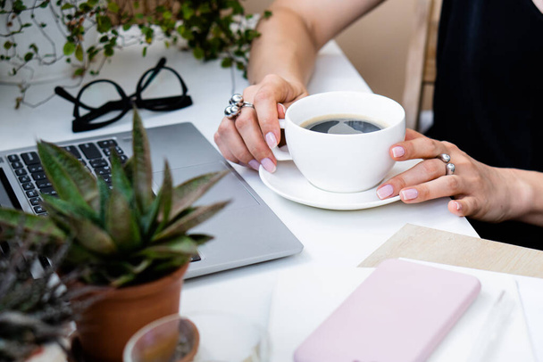 Femme main tenant tasse de café dans un bureau d'été confortable avec ordinateur portable et plantes vertes - Photo, image